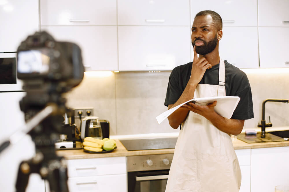 African-american bearded man smiling and reading a cooking book. Blogger shooting video for cooking vlog in kitchen at home. Man wearing an apron.